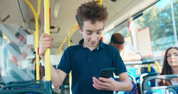 Młody chłopiec w wieku szkolnym jeździ autobusem komunikacji miejskiej do szkoły podstawowej trzymając telefon w ręku, wygląda — Wideo stockowe