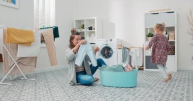 Banyo zemininde oturan bir kadın rengarenk çamaşırlarla dolu bir kasenin yanında, annesinin yanında dikkat çekmek isteyen küçük bir kız koşuyor.