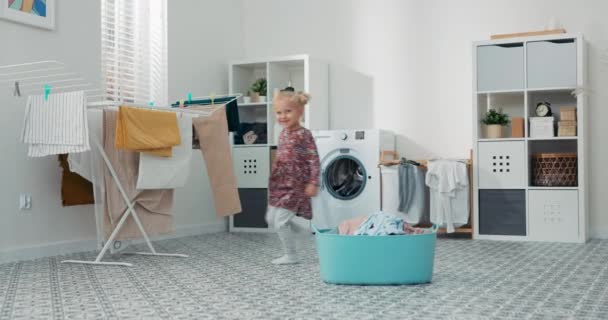 Carino adorabile bambina in abito floreale gioca in lavanderia, in bagno, il bambino corre intorno alla ciotola di vestiti accanto alla lavatrice, annoiato a casa — Video Stock