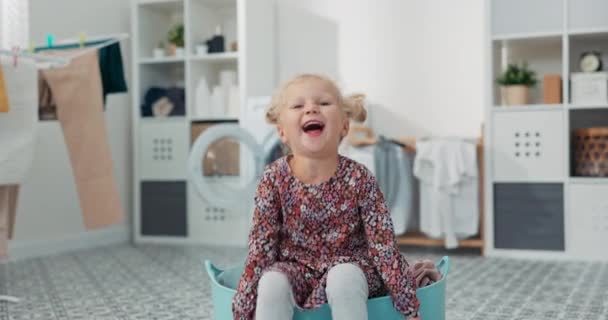 Piękna dziewczynka z blond włosami i niebieskimi oczami w kolorowej sukience siedzi w misce z ubraniami do prania, dziecko bawi się w łazience w pralni obok pralki — Wideo stockowe