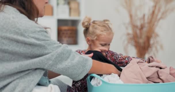 A woman and little daughter with blonde hair and blue eyes fold clean laundry taken from the clothes dryer, the little girl pulls out a sweatshirt and hands it to her mother, they watch — Stock Video
