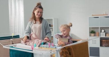 Kadın evde ev işlerini yapıyor, çamaşır odasında temiz çamaşırları kurutucuya asıyor, çocuk eğleniyor, elinde bir sepet klip tutuyor, annesiyle vakit geçiriyor.