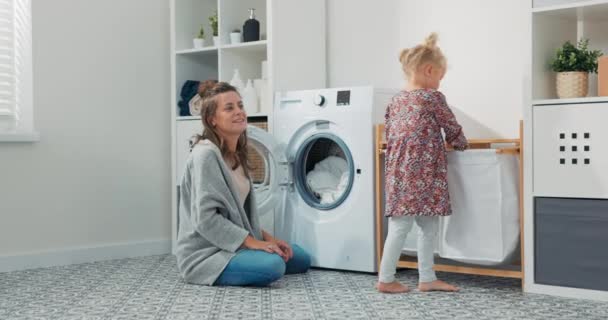Smiling mom with sweet daughter sits in bathroom, laundry girls do household chores, child hands the woman clothes from the basket to put them in the drum of the washing machine. Stock Video Footage by ©Trzykropy 