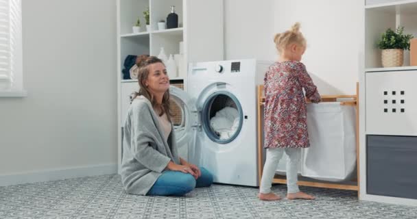 Smiling mom with sweet daughter sits in bathroom, laundry girls do household chores, child hands the woman clothes from the basket to put them in the drum of the washing machine. — Stock Video © Trzykropy #563470004