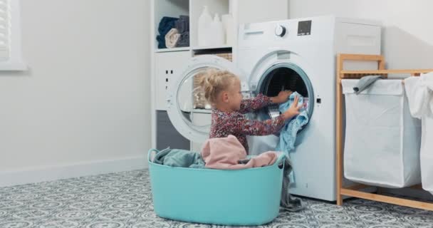 可爱的小女孩，金发，小孩坐在浴室里，一碗又一碗的衣服，女儿把东西扔进洗衣机，帮妈妈做日常事务 — 图库视频影像