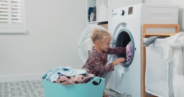 Sweet pretty little girl with blonde hair, small child sits in bathroom, laundry room by large bowl full of clothes, daughter throws things into washing machine, helps mom with daily chores — Stock Video