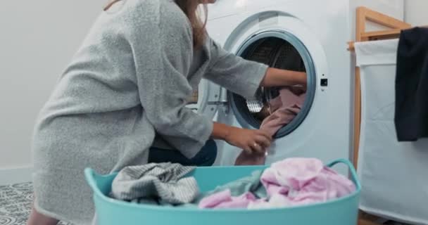 Женщина выполняет домашние обязанности в ванной комнате, прачечной, на коленях с плетеной корзиной, наполненной одеждой у стиральной машины, загружает красочные вещи в барабан — стоковое видео