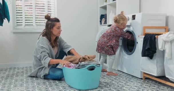 Мама с маленькой дочерью сидит в ванной комнате, они упаковывают белье в стиральную машину, смешная милая девушка кладет красочные рубашки в открытый барабан смеется торчащий язык — стоковое видео