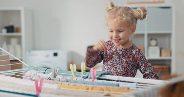 Une joyeuse petite fille en robe florale passe du temps dans la buanderie. L'enfant tient un panier de pinces à linge, qu'elle prend et attache aux serviettes de chemise suspendues au sèche-linge — Video