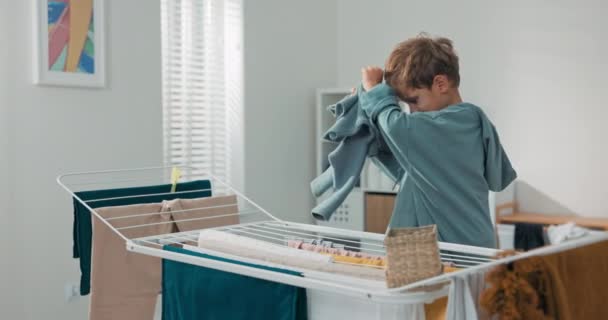 Un operoso ragazzino sorridente aiuta sua madre in lavanderia con le faccende domestiche. Il bambino tira fuori i vestiti dall'asciugatrice, li piega in una ciotola — Video Stock