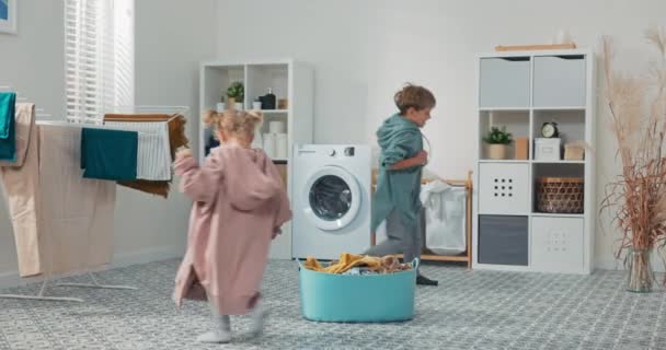 Siblings are running around playing in the laundry room, in bathroom around clothes bowl and washing machine, children are wearing oversized sweatshirts pulled from the dryer — ストック動画