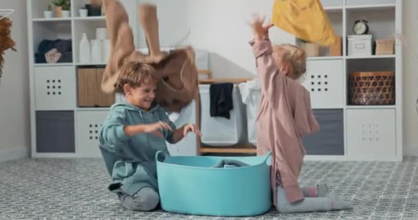 Irmãos sorridentes sentados no chão do banheiro com uma tigela cheia de lavanderia, irmã e irmão brincando, jogando roupas acima de suas cabeças — Vídeo de Stock