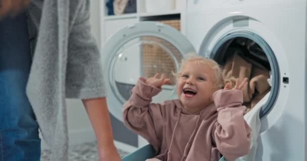 Schattig blauw ogend meisje met blond haar vastgebonden in twee broodjes dwazen rond spelen in een kom wasgoed kleren, moeder sleept meisje rond de wasruimte — Stockvideo
