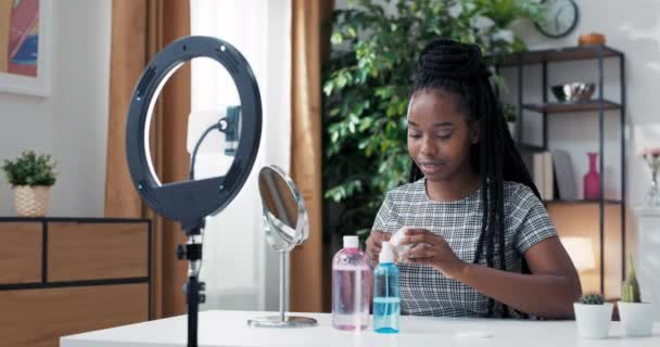 Krásná africká žena blogger ukazuje, jak používat kosmetiku. Před zrcadlem a kamerovým telefonem nahrává videonahrávku, která vysílá živě doma. Péče o pleť obličeje, aplikace hydratačního krému — Stock video