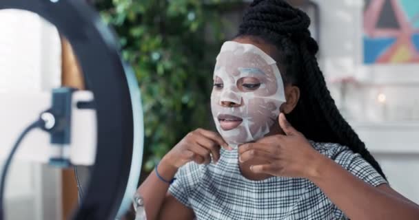 Una bella giovane donna afro-americana sta godendo di trattamenti di bellezza a casa, applicando una maschera di lenzuolo sul suo viso. Registra un tutorial per la cura della pelle per il suo canale di social media — Video Stock