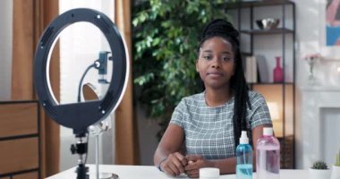 Kadın nüfuzlu kişi masasında oturmuş sosyal medya kanalı için canlı yayın yapmaya hazırlanıyor. Önünde telefon ve zil ışığı olan tripodlar. Kız kameraya gülümsüyor.