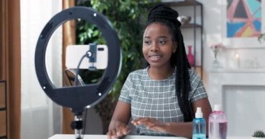 Popüler bir nüfuzlu vlogger, halka ışığı standında kurulmuş bir telefona konuşan sosyal medyada bir video yaratıyor, bir kadının önünde yüz bakım ürünleri var, kameraya bakıyor ve geniş bir şekilde gülümsüyor.