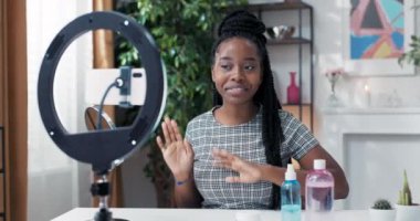 Güzellik blogcusu canlı takipçileri selamlıyor, bir lamba standında el sallıyor, kadının önünde yüz bakım ürünleri var, kameraya geniş bir gülümsemeyle bakıyor.