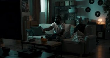 Adam gece geç saatte film izliyor, uzaktan kumandayla televizyonu kapatıyor, üstünü battaniyeyle örtüyor, yorgun adam oturma odasında kanepede uyuyor.