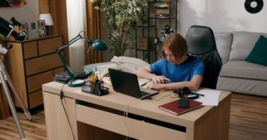 Uzun saçlı bir çocuk dizüstü bilgisayarının önünde oturup ödev yapıyor, internetten bilim videoları izliyor, sınava çalışıyor, çalışkan bir öğrenci..