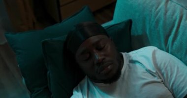 Yastıkların üzerinde rahat bir şekilde uzanan bir adamın en iyi pozu. Beyaz tişörtlü ve kafasında siyah bir mendil olan çocuk dinleniyor ve geceleri fenerlerle yanan sokağa bakıyor..