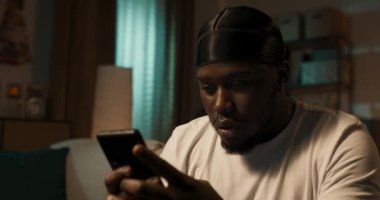 Akıllı telefon tutan, mesajlaşan ya da oyun oynayan, evdeki koltukta oturan genç Afro-Amerikalı adam, mesajları internet sitelerine göndermek için sosyal medya uygulaması kullanıyor..