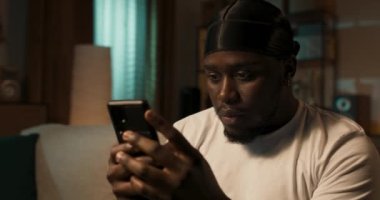 Heyecanlı Afro-Amerikalı adam oturma odasındaki kanepede dinlenirken elinde akıllı telefon tutuyor. Evde gülümseyen, iş arkadaşları ve müşterilerle internette sohbet eden mutlu bir adam. Sosyal ağlar kullanılıyor.