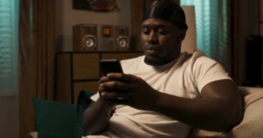 Bir adam elinde akıllı bir telefonla kanepede oturuyor, web sitelerine göz atıyor, telefona uygulamalar yerleştiriyor, elektronik alışveriş listesi yapıyor, clanedar 'ına hatırlatıcı koyuyor..