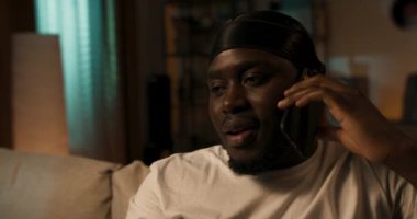 Kirli sakallı, gülümseyen adam telefonda arkadaşıyla konuşuyor, oturma odasında koltukta oturuyor ve kendini minderlere karşı dirsekle destekliyor. Arka planda duvarda vinil plaklar var.