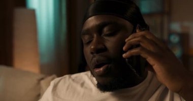 Neşeli, genç ve yakışıklı bir adam cep telefonuyla konuşuyor ve gülümsüyor, oturma odasındaki kanepede oturuyor..