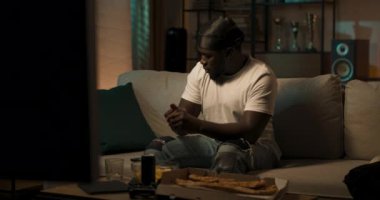 İyi akşamlar, oturma odasında. Kot pantolon ve beyaz tişört giymiş bir adam kanepede dinleniyor. Çocuk TV 'de maç izliyor, elinde bir telefon tutuyor, video sohbet yoluyla arkadaşıyla konuşuyor..