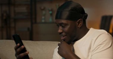 Heyecanlı Afro-Amerikalı adam oturma odasındaki kanepede dinlenirken elinde akıllı telefon tutuyor. Evde gülümseyen, iş arkadaşları ve müşterilerle internette sohbet eden mutlu bir adam. Sosyal ağlar kullanılıyor.
