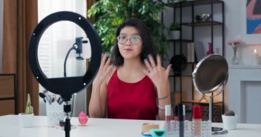 Kız vlogger, izleyiciler için makyaj videosu kaydediyor. Telefona el sallıyor. Kürsüde halka ışığıyla duruyor..