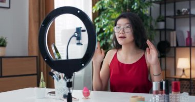 Güzellik blogcusu canlı takipçileri selamlıyor, bir lamba standında el sallıyor, kadının önünde yüz bakım ürünleri var, kameraya geniş bir gülümsemeyle bakıyor..