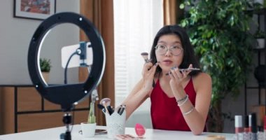 Bir kız telefon tripoduyla halka ışığı kullanarak video çekiyor. Makyaj sanatçısı yüz hatlarını nasıl düzelteceğine dair bir ders hazırlar. Yanağa brozer sürmek için fırça kullanır..