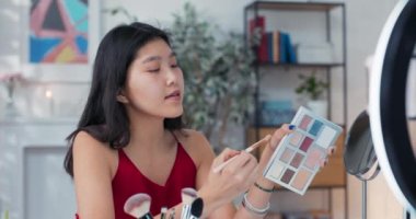 Gülümseyen esmer kadın güzellik blogcusu göz farı paleti gösteren ürünler öneriyor. Akıllı telefon ve halka ışığıyla video kaydediyor. Kadın makyaj sanatçısı etkileyicisi özel ders oluşturur.