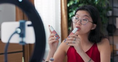 Kadın güzellik blogcusu günlük makyaj rutinini kaydediyor. Tripodun üzerinde telefon ile halka ışığı kullanıyor. Kozmetikler hakkında canlı yayın yapan koyu tenli nüfuzlu biri, iki farklı renkte ruj kullanıyor..