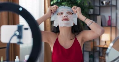Güzellik blogcusu makyaj ürünlerini sosyal medyada gözden geçirmek için canlı yayında kumaş güzellik maskesi takıyor. Nüfuzlu kişi tripoda ve halka lambasında akıllı telefon kullanır..