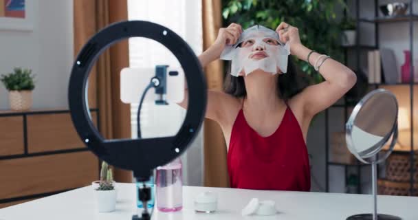 美容ブロガーは 顔にファブリック美容マスクを適用しながら ソーシャルメディア上の化粧品を確認するためのライブストリーミングです インフルエンサーは三脚とリングライトにスマートフォンを使用します — ストック動画