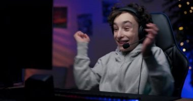 Profesyonel oyuncu monitör önünde bilgisayar oyunları oynayarak vakit geçirir, çocuk el çırpar, kulaklığı takar ve neon ışıklarla aydınlatılmış takım odasıyla şakalaşır..