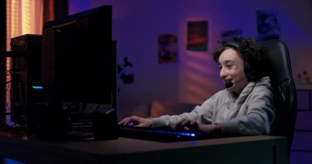 ヘッドセットと小さなゲーマー 少年は夜遅くにコンピュータモニタの前に座っています 彼のチームとマルチプレイヤーシューティングゲームのビデオゲームをプレイ 部屋は Ledライトによって点灯 — ストック動画