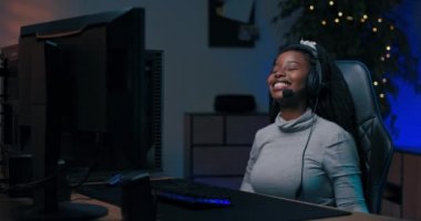 Koyu tenli profesyonel oyuncu bilgisayar başında kulaklıkla konuştuğu takımla oynuyor, takım sanal çevrimiçi şampiyonluğu kazanıyor, kız zafere sevinçle el kaldırıyor..