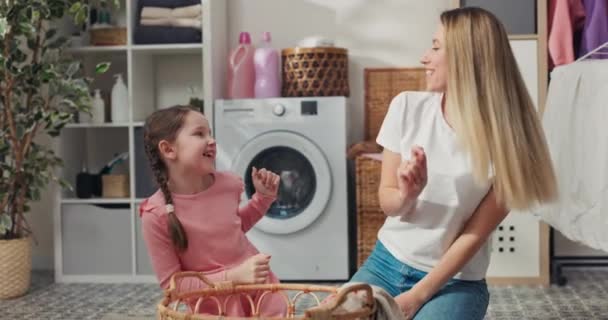 一个穿着粉色连衣裙和辫子的小女孩和妈妈坐在洗衣房的地板上 手里拿着一个柳条篮 穿着漂亮的衣服 随着收音机里的音乐在房间里跳舞 — 图库视频影像