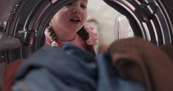 ピグテールの笑顔の女の子は洗濯機をアンパック ドラムからカラフルなぬれたきれいな香りの服を取り出し 洗濯室で時間を過ごす家事 — ストック動画