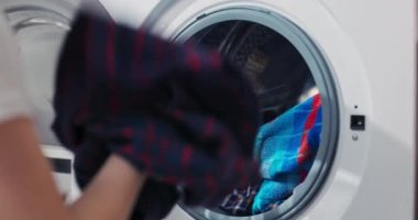 Close up shot of the drum. A little girl puts colorful clothes in the wash. The child helps mother with the housework, closes the washing machine door.