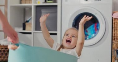 Elbise kâsesinde oturan sevimli sarışın kızın yakın çekimi. Kadın eğlence olsun diye kızı çeker anne kızıyla çamaşır odasında vakit geçirir..