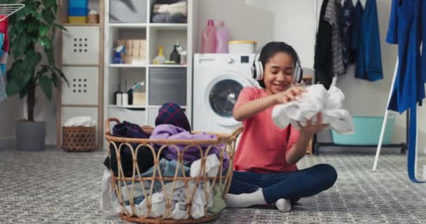 微笑的女孩坐在洗衣房的地板上 面对着一个大柳条筐 手里拿着要整理的衣服 在家务活上花时间 帮助父母 听音乐 — 图库视频影像