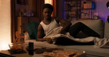 A student wakes up from an afternoon nap, sits down on the couch, reaches for the remote and turns on the TV, his favorite show.