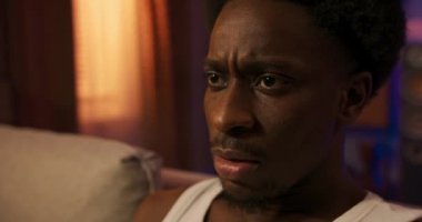 A disgusted man with a strange face sits on the sofa in a comfortable sleeveless white t-shirt, an afro guy watches TV in the afternoon, the golden sun comes through the window blind.