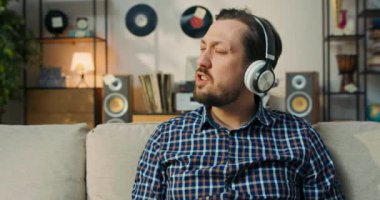 Sakallı, siyah saçlı, kablosuz kulaklıkla müzik dinleyen bir çocuğun yakın çekimi. Adam favori yabancı şarkıları söylüyor, dizlerine vuruyor ritme ayak uyduruyor..